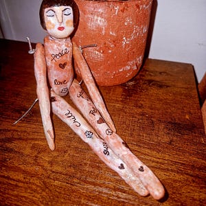 May include: A handcrafted figurine with a light brown, textured body and a dark brown bob hairstyle. The figure features hand-painted text including 'peace', 'love', 'chill', and 'good vibes', along with small heart and flower symbols. The arms and legs are elongated, and the figure is constructed with visible wire joints.