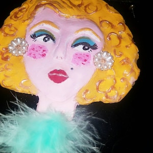May include: A hand-painted, decorative face with yellow curly hair, pink skin, and red lips. The face has blue eyeshadow, pink blush, and pearl earrings. A turquoise feather boa adorns the neck.