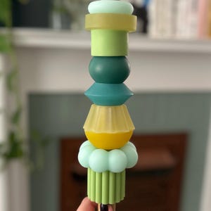 Puede incluir: Una varilla de madera con una pila de formas geométricas de colores. Las formas están hechas de un material liso y gomoso e incluyen los colores verde, amarillo y azul.