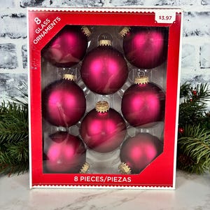May include: A box containing 8 round glass ornaments in a deep magenta color with a matte finish. Each ornament features a gold-colored metal cap at the top. The packaging is red with white text that reads 8 GLASS ORNAMENTS and 8 PIECES/PIEZAS.