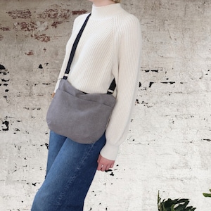 May include: A grey canvas crossbody bag with a black adjustable strap. The bag is being worn by a person wearing a white sweater and blue jeans.