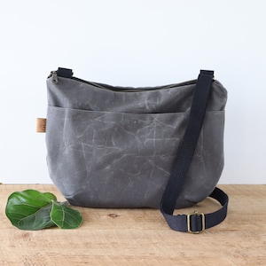 Sac à bandoulière arrondi en toile cirée pour femme et homme, avec fermeture éclair sur le dessus et poche extérieure.Sac robuste en toile cirée pour l'extérieur et les voyages.