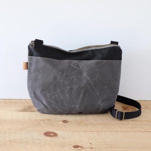 Black and Charcoal rounded waxed canvas crossbody bag, with top zipper and exterior pocket. Rugged Waxed Canvas Bag for outdoors and travel.