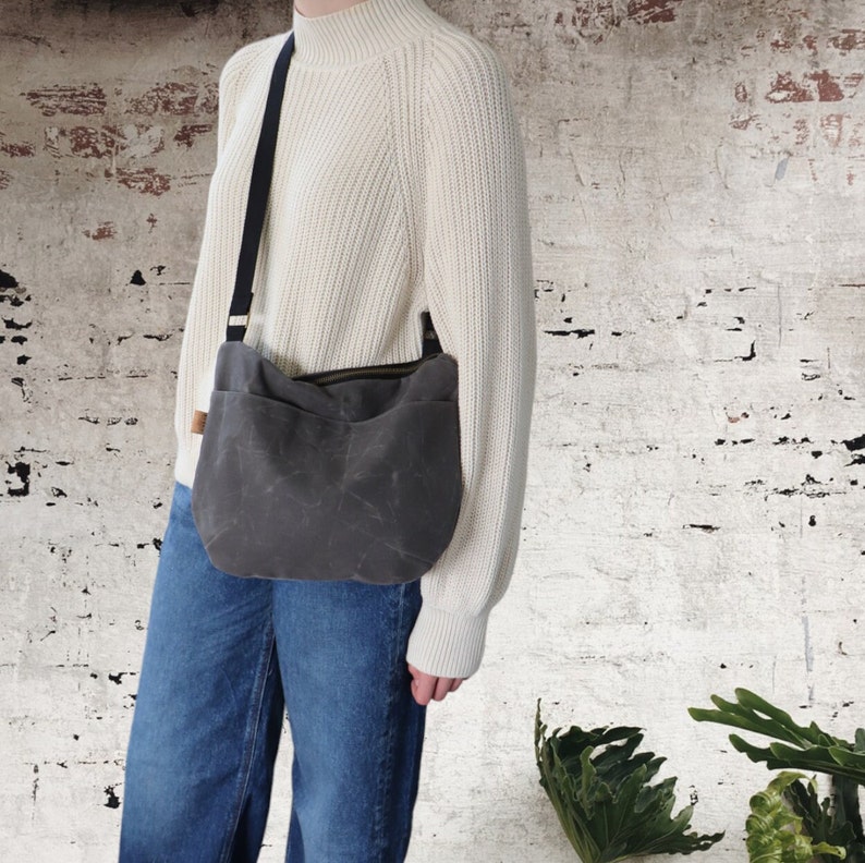 May include: A gray canvas crossbody bag with a black strap. The bag has a flap with a zipper closure and a front pocket.