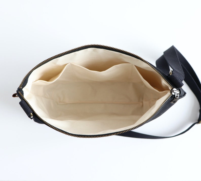 May include: Black canvas crossbody bag with a zipper closure and a beige fabric interior. The bag has two interior pockets.