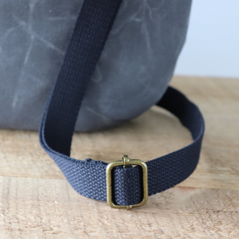 May include: A close-up of a dark gray canvas bag with a black adjustable strap and a brass buckle.