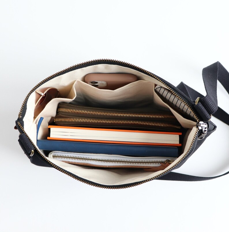 May include: A black canvas crossbody bag with a zipper closure and multiple interior pockets. The bag is open and filled with various items, including a phone, books, and a wallet.