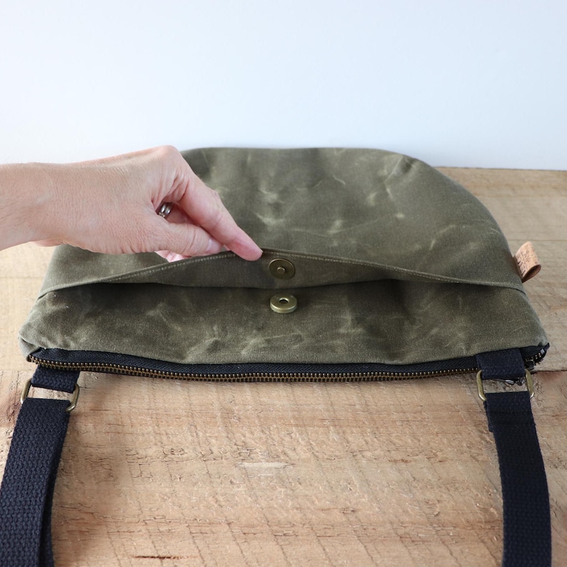 May include: A green canvas crossbody bag with a zipper closure and a magnetic snap closure. The bag has a black adjustable strap.