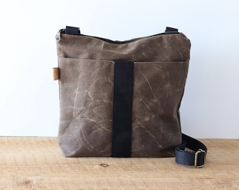 May include: A brown waxed canvas crossbody bag with a black strap and a zipper closure. The bag has a worn look and a small pocket on the front.