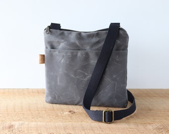 May include: A gray canvas crossbody bag with a black adjustable strap and a zipper closure. The bag has a front pocket and a small leather tag.