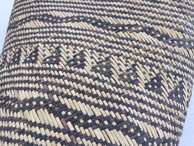 May include: Close-up of a woven basket with a black and natural straw pattern. The basket is made of natural materials and has a unique, intricate design.