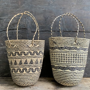 May include: Two woven tote bags with black and natural colour patterns. Each bag has a rounded shape with a handle. One bag features a zigzag and triangle design, while the other has a diamond and wavy pattern. The bags are set against a dark wood background.