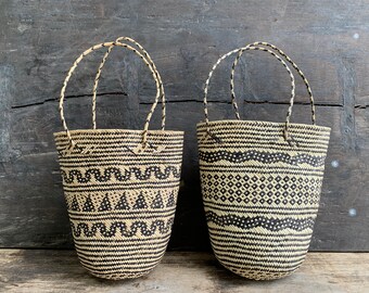 Puede incluir: Dos bolsas de mano tejidas con patrones negros y naturales. Cada bolsa tiene forma redondeada con asa. Una bolsa presenta un diseño en zigzag y triángulo, mientras que la otra tiene un patrón de diamantes y ondulado. Las bolsas están sobre un fondo de madera oscura.