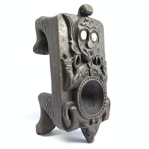 May include: A dark wood carving of a mythical creature with a round hollow in the center. The creature has a human-like face with large eyes and a wide mouth. The carving is on four legs and has intricate details.