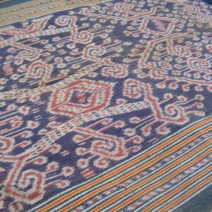 Puede incluir: Un primer plano de una tela tejida con un complejo patrón geométrico en tonos de rojo, blanco y azul sobre un fondo azul oscuro. La tela tiene una superficie texturizada y un borde con flecos.