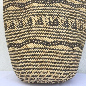 May include: A large, woven basket with a black and natural straw pattern. The basket is made of natural materials and has a rounded shape.