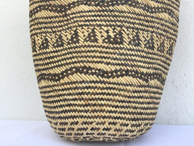 May include: A large, woven basket with a black and natural straw pattern. The basket is made of natural materials and has a rounded shape.