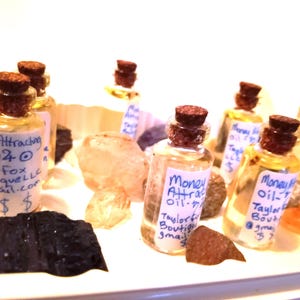 Puede incluir: Pequeñas botellas de vidrio con tapones de corcho, etiquetadas como "Money Attracting Oil" y rodeadas de cristales y piedras. Las botellas tienen etiquetas manuscritas con la información del vendedor y símbolos de dólar.