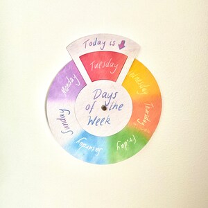 May include: A circular days of the week chart featuring a rainbow color gradient. The center is white with the text "Days of the Week" and a metal fastener. A movable top section displays "Today is" with a purple arrow pointing to Tuesday.