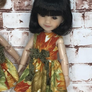 May include: A doll wearing a fall-themed dress with a leaf pattern in shades of orange, green, and gold. The dress has a green bow at the waist and a white lace trim at the hem. The doll has black hair.