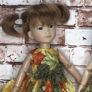 May include: A doll wearing a yellow, green and orange dress with a green bow and white lace trim. The dress has a leaf pattern.