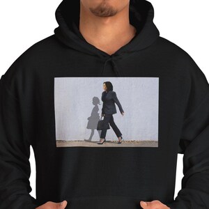 May include: Black hooded sweatshirt with a front graphic. The graphic is a photograph of a person in a dark suit walking in front of a white wall, with their shadow visible.