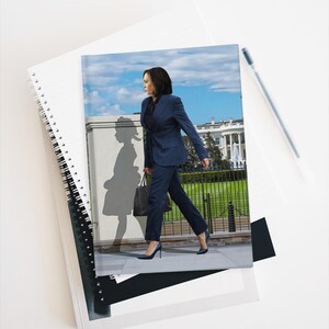 May include: A spiral-bound notebook with a cover featuring an image of a person in a navy blue suit walking in front of the White House. A pen rests on the white surface next to the notebook.