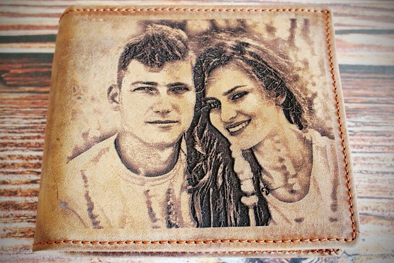 Puede incluir: Una cartera de cuero marr&oacute;n claro con un retrato grabado con l&aacute;ser de una pareja sonriente. La cartera tiene un borde cosido con hilo naranja en contraste. La imagen es un retrato en tonos sepia de un hombre y una mujer.