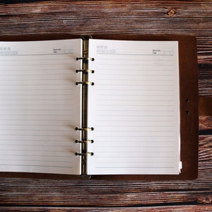 May include: Open brown leather journal with lined white paper pages. The journal is open, revealing the blank pages, and is set against a dark wooden background. The journal has a metal ring binder.