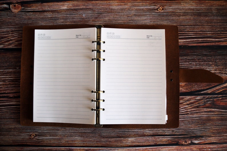 May include: Open brown leather journal with lined white paper pages. The journal is open, revealing the blank pages, and is set against a dark wooden background. The journal has a metal ring binder.