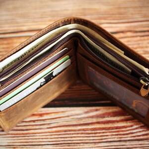 Puede incluir: Cartera de cuero marr&oacute;n abierta, llena de efectivo, tarjetas de cr&eacute;dito y tarjetas de identificaci&oacute;n. La cartera tiene un cierre de cremallera y m&uacute;ltiples compartimentos. El cuero tiene un aspecto envejecido.
