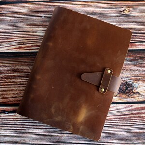 May include: A brown leather journal with a strap closure. The journal is rectangular and has a textured surface. The strap is secured with two brass-colored rivets. The journal is lying on a wooden surface.