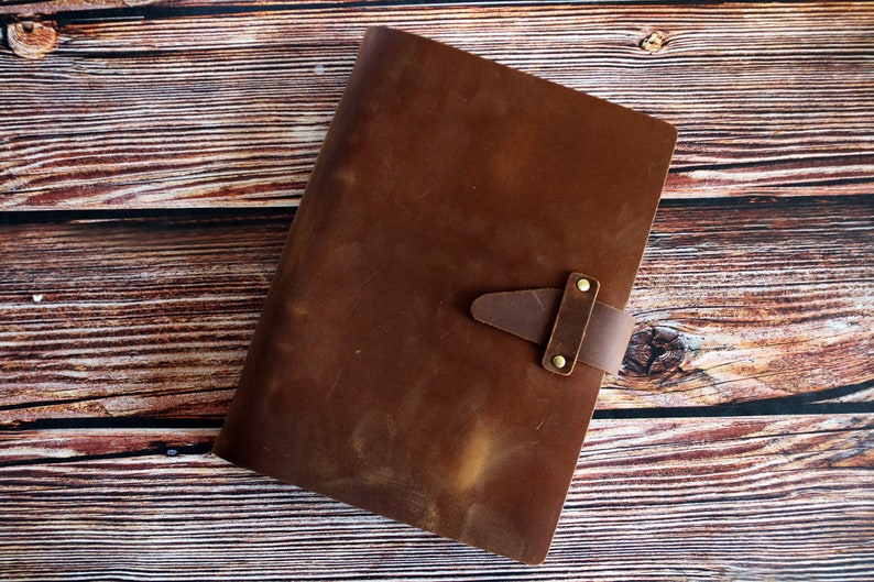 May include: A brown leather journal with a strap closure. The journal is rectangular and has a textured surface. The strap is secured with two brass-colored rivets. The journal is lying on a wooden surface.