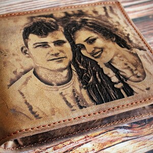 Puede incluir: Cartera de cuero marr&oacute;n con un retrato grabado de una pareja. La cartera est&aacute; cerrada, mostrando la imagen. Los bordes est&aacute;n cosidos con un hilo en contraste, con un dise&ntilde;o r&uacute;stico.