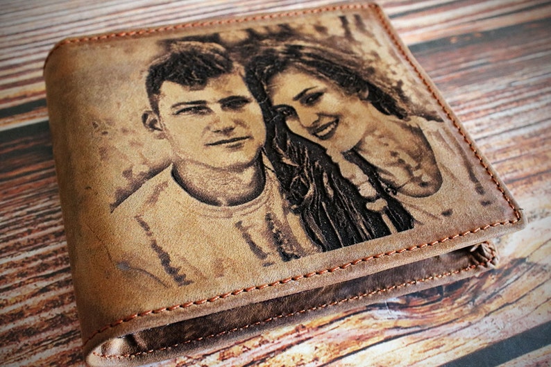 Puede incluir: Cartera de cuero marr&oacute;n con un retrato grabado de una pareja. La cartera est&aacute; cerrada, mostrando la imagen. Los bordes est&aacute;n cosidos con un hilo en contraste, con un dise&ntilde;o r&uacute;stico.