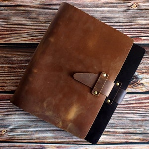 May include: A brown leather journal with a strap closure and brass rivets. The journal has a distressed appearance and a black interior. The cover is a rich, warm brown color, and the journal is set against a wooden background.