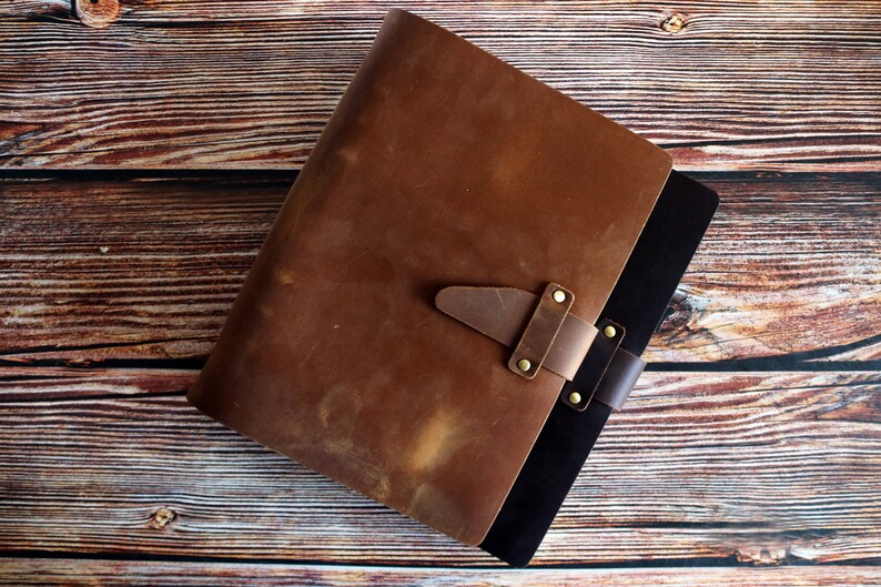 May include: A brown leather journal with a strap closure and brass rivets. The journal has a distressed appearance and a black interior. The cover is a rich, warm brown color, and the journal is set against a wooden background.