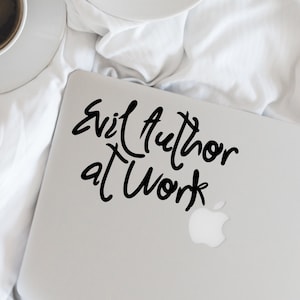 May include: A white laptop with a black sticker that says "Evil Author at Work". The laptop is on a white bedspread with a cup of coffee and a dessert on a plate.