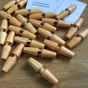 May include: A collection of light brown wooden toggle buttons, each featuring a smooth, cylindrical shape with a central groove and a single hole for attachment. The buttons are scattered, showing their natural wood grain and polished finish.