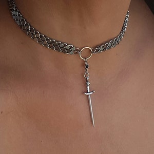 May include: A silver chain choker necklace with a dangling sword pendant. The sword has a black stone at the top.