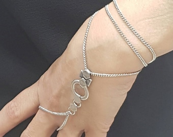 May include: A silver chain bracelet with a decorative charm that connects to a finger ring. The bracelet is wrapped around the wrist twice.