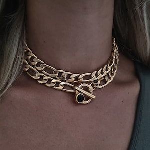 Op de afbeelding: Een gouden ketting met een zwarte stenen toggle sluiting. De ketting is gelaagd en heeft een chunky, curb chain design.