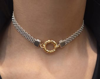 May include: A silver and gold choker necklace. The necklace features a silver chain with a gold-colored circular centerpiece. The necklace is worn on a person wearing a black shirt.