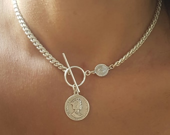 May include: A silver chain necklace with a toggle clasp and a coin pendant featuring a profile of a woman. The coin is attached to the chain with a smaller round charm.