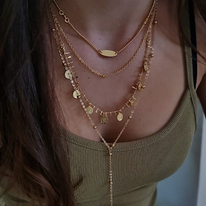 May include: A layered gold necklace with a long pendant chain and small coin-shaped charms. The necklace is worn by a person with long brown hair.