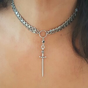 May include: A silver chain choker necklace with a small sword pendant. The chain is thick and has a curb link design. The sword pendant is detailed and has a black stone at the top.