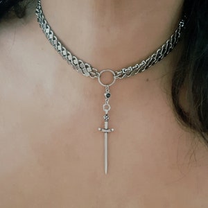 May include: A silver chain choker necklace with a small sword pendant. The chain is made of interlocking links and the sword has a black gemstone at the top.