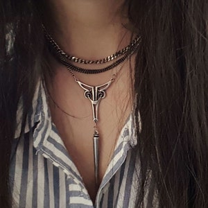 May include: A silver necklace with a long, pointed pendant and a layered chain design. The necklace is worn by a person with long, dark hair.