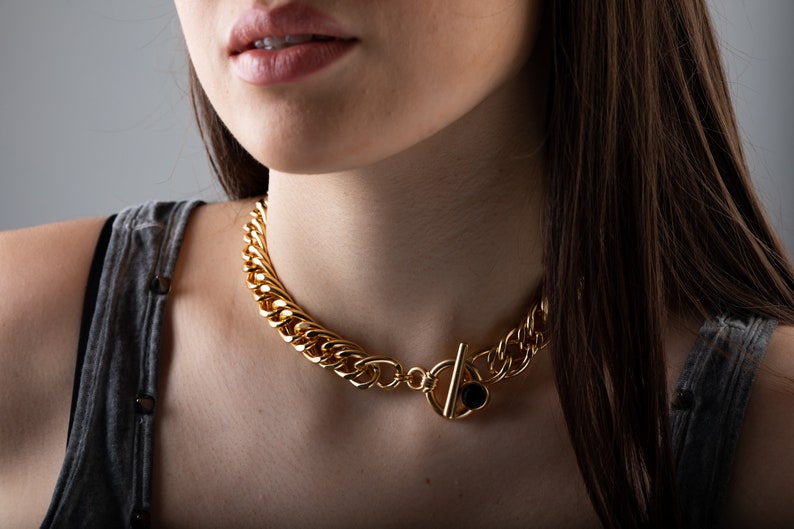 May include: A gold chain necklace with a large, chunky link design. The necklace has a black stone accent on the clasp.