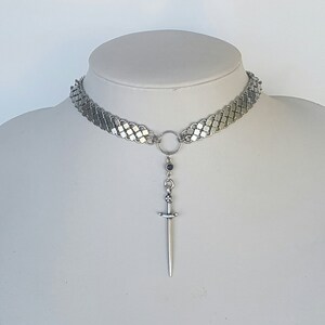 May include: A silver chain choker necklace with a sword pendant. The chain is made of small, interlocking squares. The sword pendant has a black gemstone at the top.